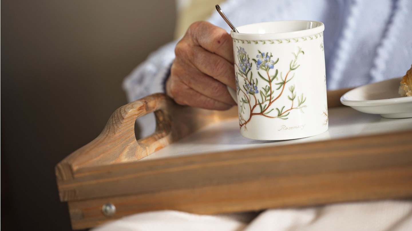 Häusliche Pflege mit Heilpflanzen und Hausmitteln: Fotoausschnitt eines Pflegebedürftigen, der im Bett ein Tablett mit einer großen Tasse Kräutertee hält.