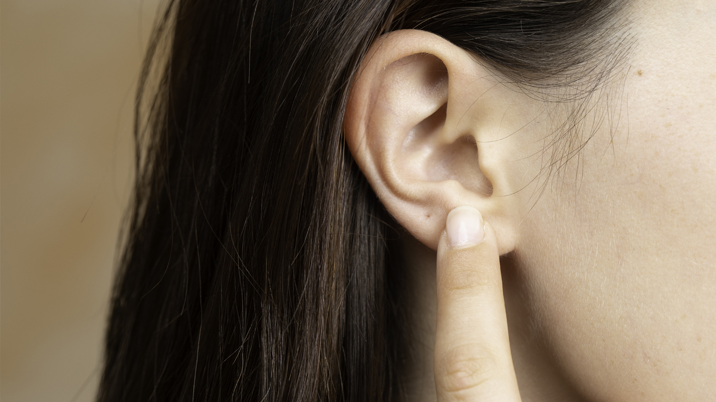 Ohren reinigen: Ein Zeigefinger zeigt auf das Ohr einer Frau mit braunen Haaren.
