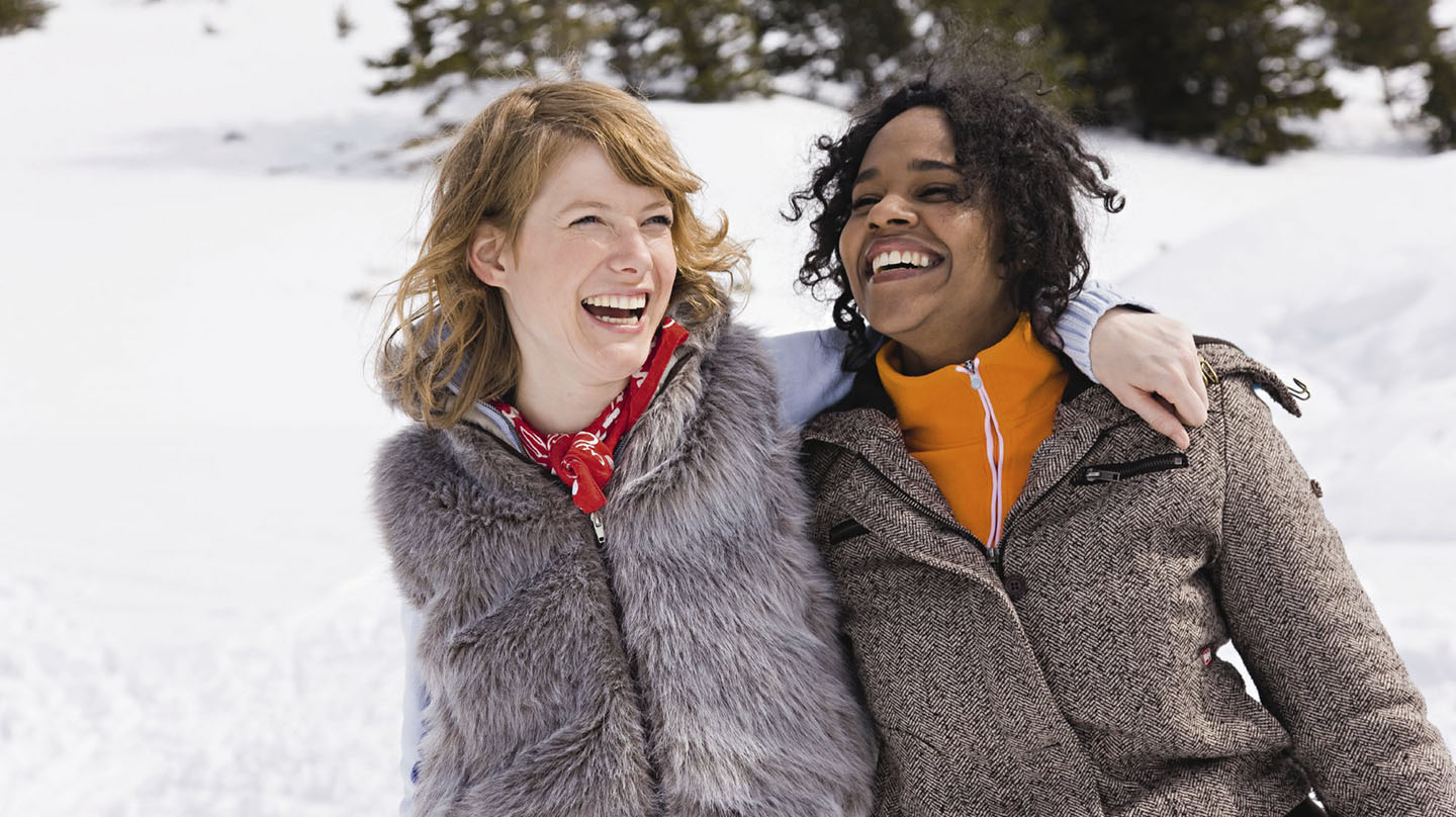 Vitamin D Mangel vermeiden: Zwei junge lachende Frauen in Wintermänteln gehen in verschneiter Landschaft spazieren.